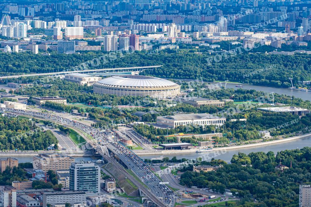 На фотографии представлен вид на Лужники, один из крупнейших спортивных комплексов в Москве, расположенный на берегу Москвы-реки. На заднем плане видны жилые районы и зелёные насаждения городского парка. Фотография подходит для использования в спортивных публикациях, материалах о городском планировании и туристических проспектах, а также может служить иллюстрацией в статьях о важных городских достопримечательностях.