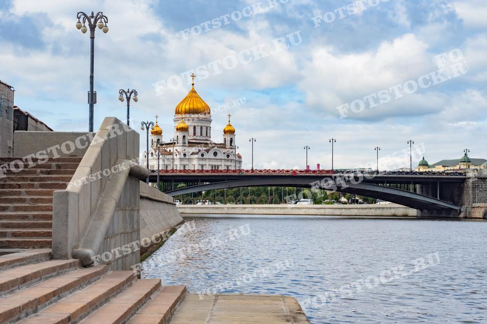На фотографии изображен Храм Христа Спасителя на фоне Москвы-реки. Впереди видны ступени городской набережной и пешеходный мост. Летний день с облачным небом создает спокойную атмосферу.