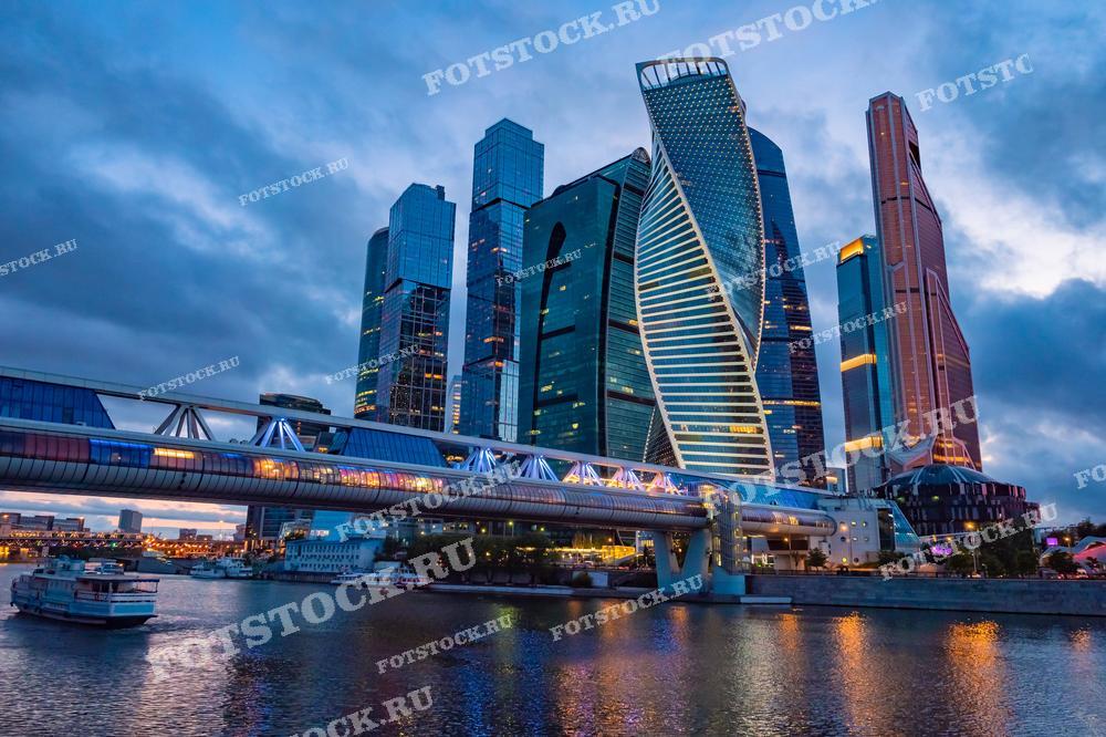 На фотографии изображены высотные здания делового центра Москва-Сити в вечернее время. Слева видно подсвеченный пешеходный мост "Багратион", соединяющий две части комплекса. Небоскрёбы отражаются в водах реки Москва, добавляя глубины и динамики городской панораме. Вечерние огни и облачное небо создают контрастный и атмосферный фон для современного делового района Москвы. Это один из символов современной столицы.