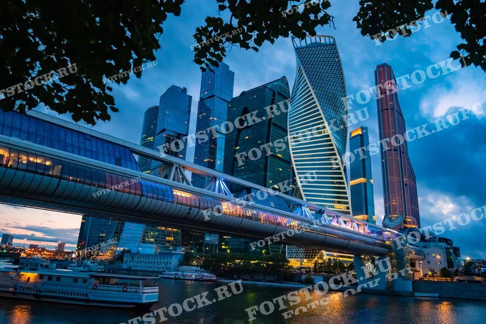 На фотографии изображен вечерний вид на небоскребы делового центра Москва-Сити и мост "Багратион". Закатное небо и освещение мостов и зданий создают впечатляющий образ современного мегаполиса.