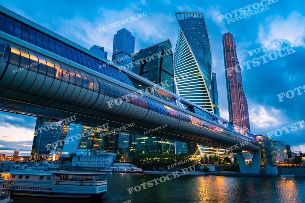 На фотографии изображен вечерний вид на деловой центр Москва-сити с мостом "Багратион". Современные небоскребы и подсвеченный мост создают впечатляющий городской пейзаж. Внизу видна Москва-река с прогулочными теплоходами.