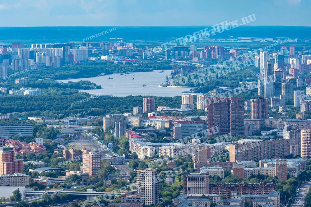 На фотографии изображён панорамный вид Москвы, снятый с большой высоты. На переднем плане видны многоэтажные жилые дома и зелёные насаждения, а на заднем плане — далекие районы города и река Москва, по которой плавают корабли. Это изображение иллюстрирует современную застройку и обширные пространства мегаполиса. Такие фото могут быть использованы для статей и исследований о градостроительстве, публикаций о Москве, а также в качестве фонов для дизайна и рекламы.