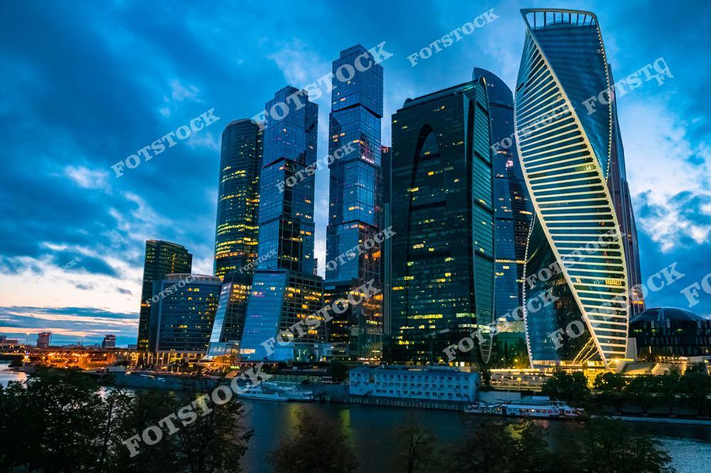 На фотографии запечатлены небоскрёбы делового центра Москва-Сити в вечернее время. Башни "Эволюция", "Федерация" и другие отражают огни города в стеклянных фасадах, подчёркивая динамику и современность Москвы. На переднем плане видна Москва-река с причалами и прогулочными судами.