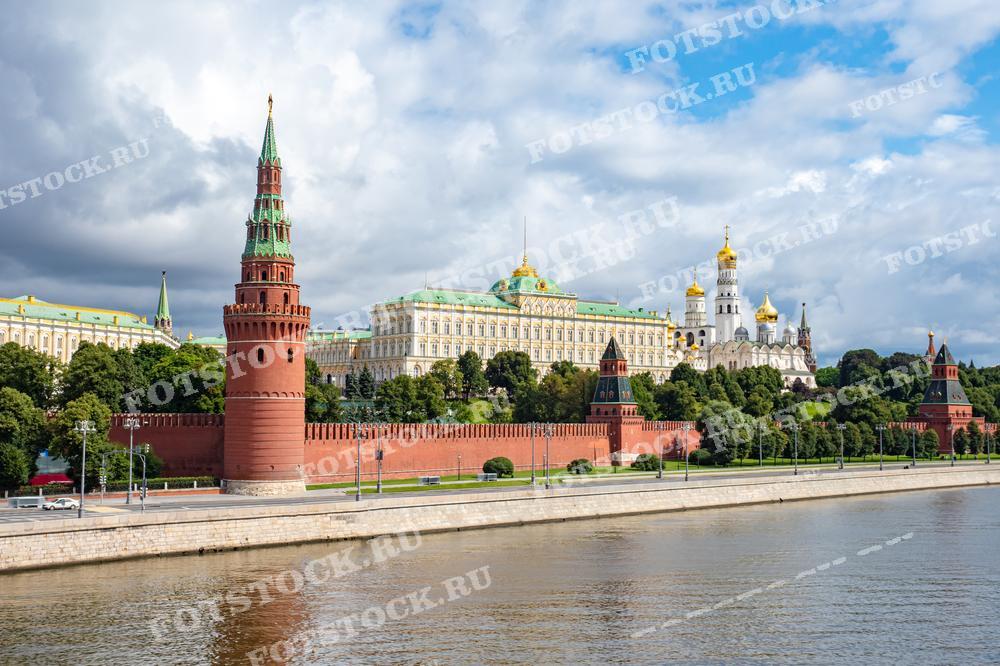 На фотографии изображен летний вид на Кремль и Москву-реку. Красные башни Кремля и золотые купола Большого Кремлевского дворца выделяются на фоне зеленых деревьев и облачного неба. Этот вид является визитной карточкой столицы Российской Федерации.