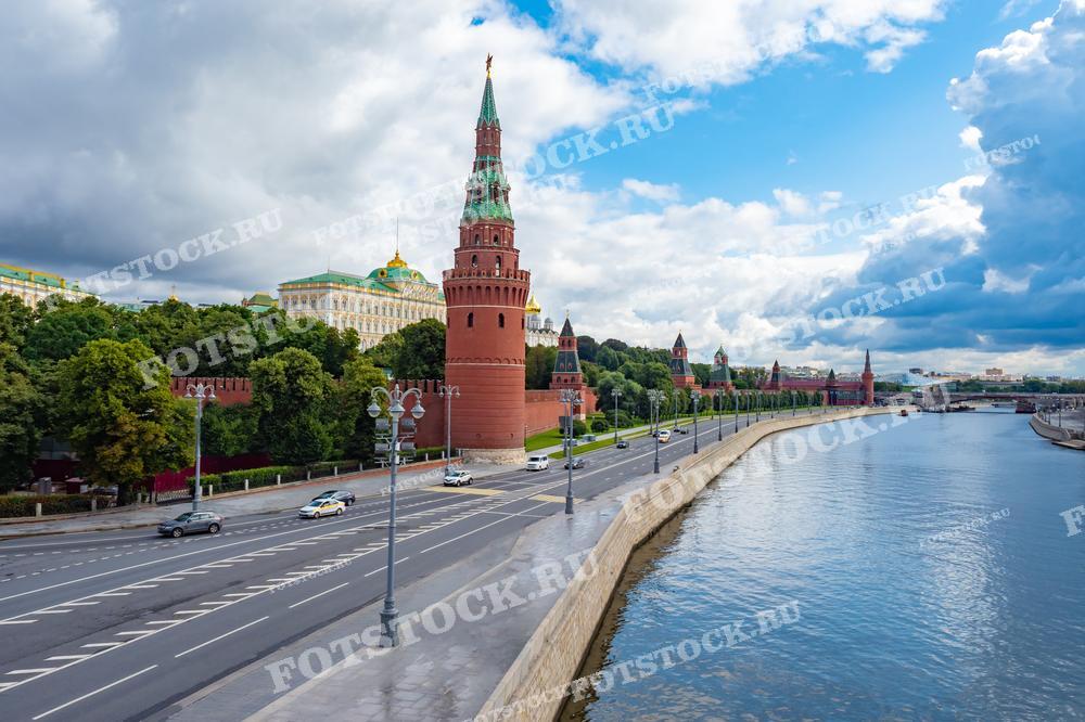 На фотографии представлен вид на Московский Кремль и Кремлёвскую набережную. Видно, как Кремлёвские стены эффектно обрамляют комплекс зданий, а за ними возвышается зелёный лесопарк. Дорога вдоль набережной показывает динамику городской жизни с движущимися автомобилями. Фотография может быть использована для учебных материалов по истории и архитектуре, в туристических брошюрах и как декоративный элемент в оформлении офисов и жилых помещений.