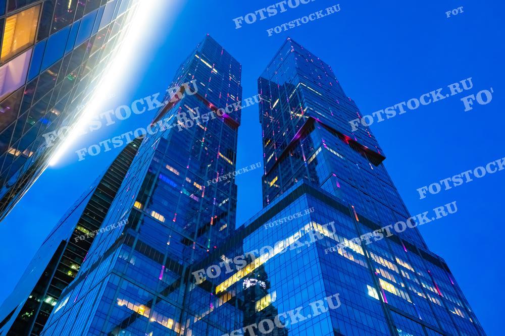На фотографии изображены небоскребы Москва-Сити, сияющие в вечернем свете. Стеклянные фасады зданий отражают небо, подсвеченные разноцветными огнями.