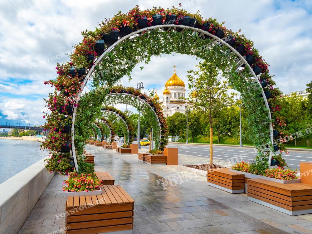 На фотографии изображена набережная в центре Москвы, украшенная цветочными арками. Вдали виднеется золотой купол Храма Христа Спасителя, одной из главных святынь России. Пешеходная зона оборудована скамейками и зелеными насаждениями, создающими уютную атмосферу для прогулок.