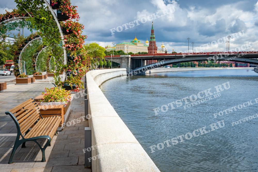 На фотографии изображена набережная Москвы-реки, украшенная цветочными арками и лавочками для отдыха. На заднем плане виден один из мостов через реку, а за ним — стены Кремля с его величественными башнями и куполами. Справа возвышается Большой Кремлевский дворец, который дополняет пейзаж исторической Москвы. Яркие цветы, свежая зелень и река создают атмосферу спокойствия и умиротворения в центре большого города.