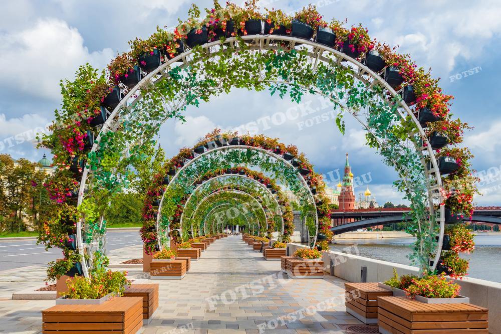 На фотографии изображены цветочные арки на набережной Москвы-реки. Летний день подчеркивает яркие краски цветов и зелени. На заднем плане виден Кремль, создавая живописный и привлекательный вид для прогулок вдоль реки.