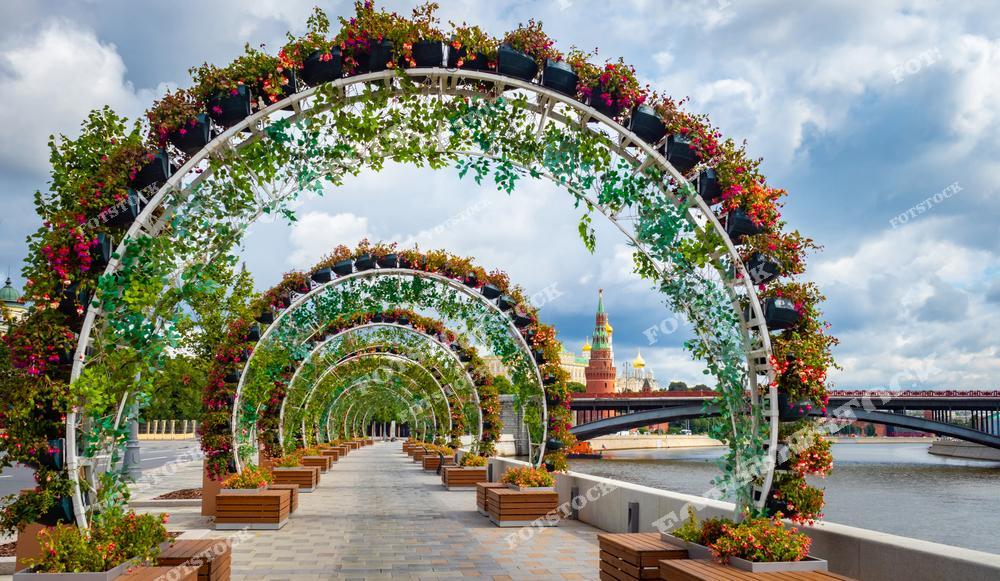 На фото изображен красочный пешеходный мост, украшенный серией арочных конструкций, обвитых зелёными растениями и цветами. Мост ведёт к кремлёвской стене и башне, видны облака и синее небо. Вдоль моста расставлены деревянные скамейки и урны, создающие уютную атмосферу для прогулок по Москве.