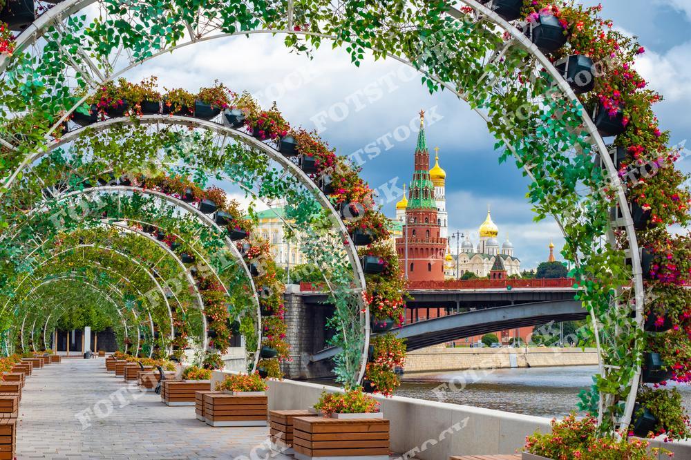 На фотографии изображена благоустроенная прогулочная зона с цветочными арками у набережной Москвы-реки. В центре кадра виднеются Спасская башня Кремля и золотые купола соборов. Арки, украшенные зеленью и яркими цветами, создают уютную атмосферу и подчёркивают красоту городской природы. Прогулочная зона оборудована лавочками и идеально подходит для отдыха. Летний день с мягкими облаками на небе добавляет снимку яркости и гармонии.