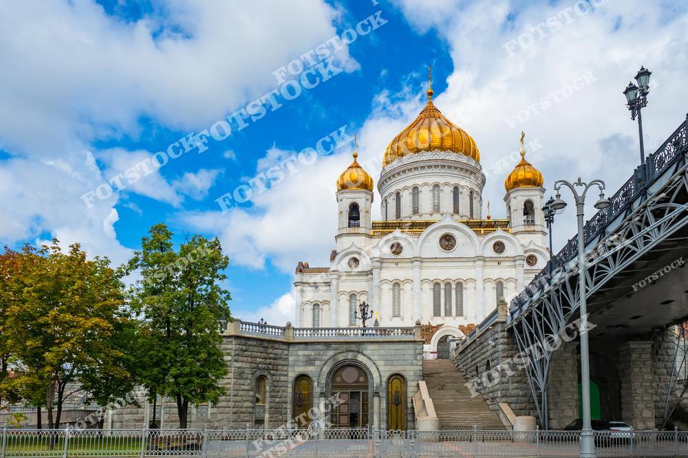 На фотографии изображён Храм Христа Спасителя, одна из главных религиозных и архитектурных достопримечательностей Москвы. Белоснежные стены храма с золотыми куполами прекрасно контрастируют с голубым небом и зеленью деревьев. Этот православный храм был воссоздан в 1990-х годах на месте разрушенного здания и сегодня является символом духовной и исторической связи России. На переднем плане виден благоустроенный парк, а сбоку — мост с элегантными металлическими перилами. Светлый солнечный день подчеркивает красоту и величие этого места.
