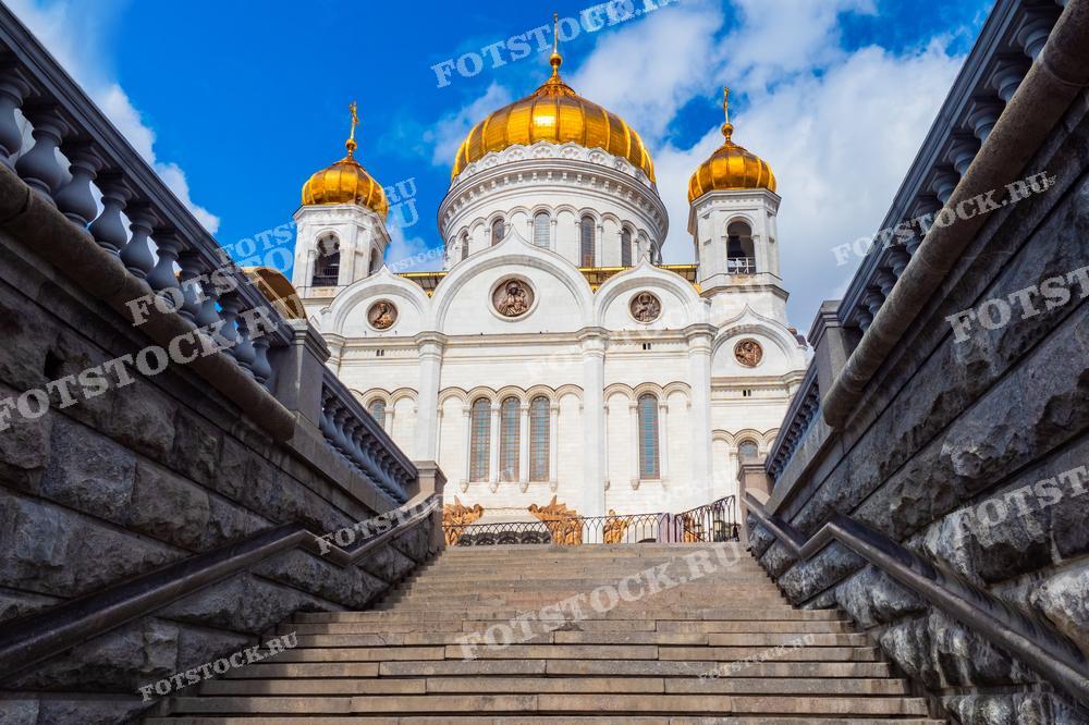 На фотографии изображен Храм Христа Спасителя, снятый с лестницы, ведущей к храму. Золотые купола ярко сверкают на фоне летнего голубого неба с облаками. Каменные перила и ступени подчеркивают величественность архитектуры храма.