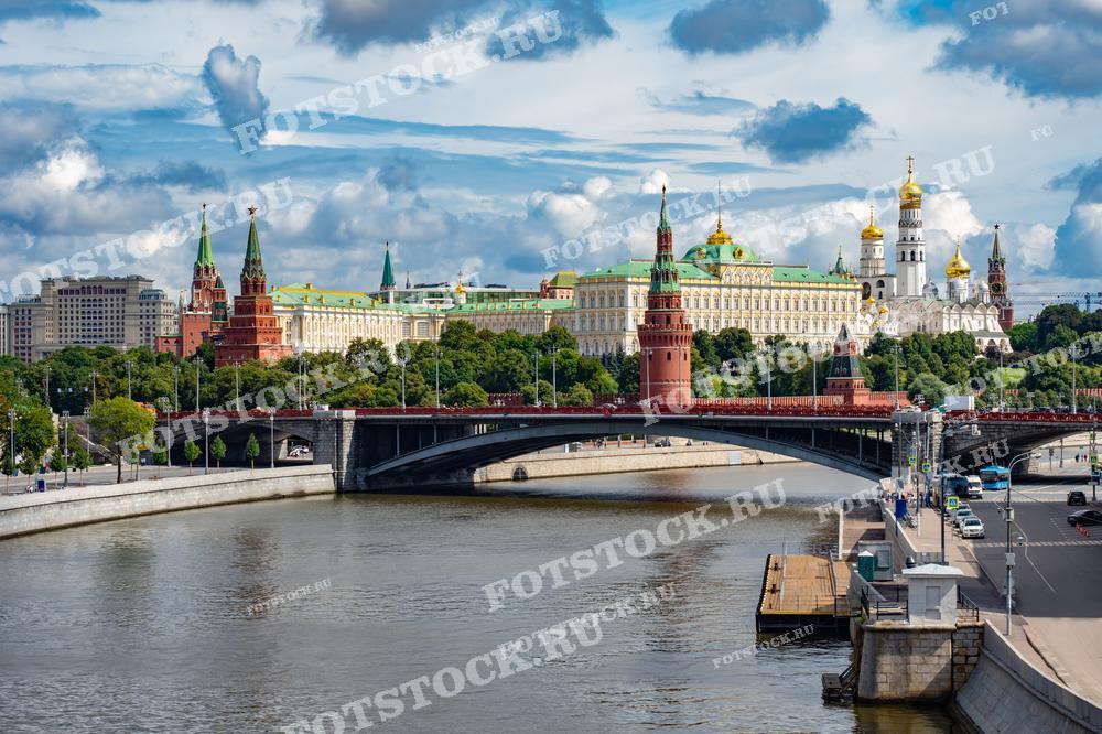 На фотографии изображены знаменитые стены Московского Кремля, включая несколько его башен, а также Большой Кремлевский мост, пересекающий Москву-реку. На заднем плане видны купола соборов и Большой Кремлевский дворец, символы российской государственности. Яркие летние краски подчеркивают зелень парков, окружающих Кремль. Мост и река Москва добавляют городской динамики этому историческому месту. Фото отражает гармонию природы и архитектуры в сердце столицы.