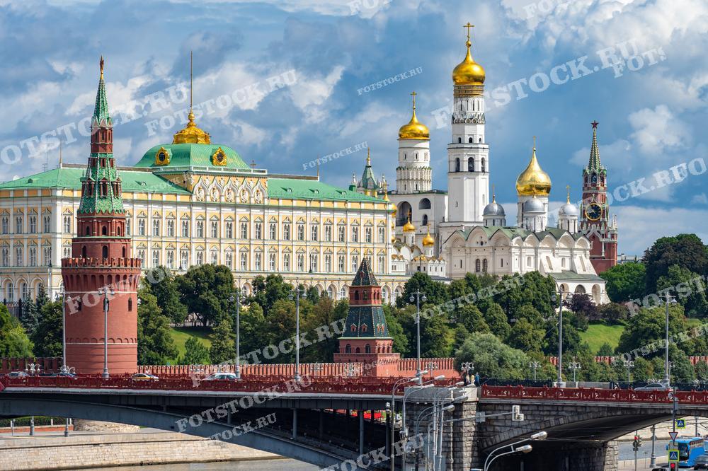 На фотографии запечатлён вид на Московский Кремль, включая Большой Кремлёвский дворец, колокольню Ивана Великого, Успенский собор и Спасскую башню. Фото показывает изысканность и детализацию архитектурных элементов, а также яркие золотые купола, которые характерны для русского архитектурного стиля. Зелёные насаждения и фон облачного неба придают изображению особую выразительность и красоту. Это изображение отлично подойдёт для использования в образовательных и культурных публикациях, туристических материалах и книгах по истории.
