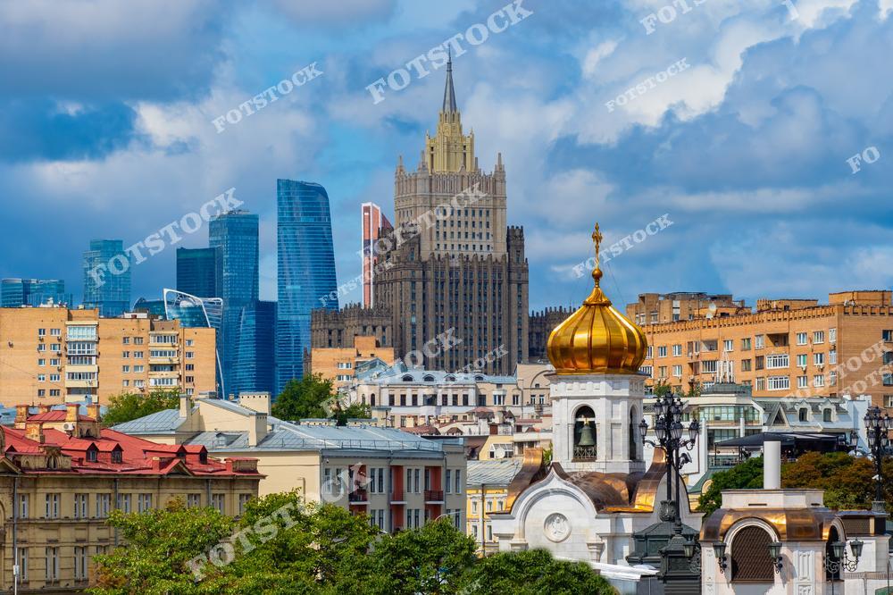 На переднем плане виден золотой купол одного из московских храмов, символизирующий богатое духовное наследие города. На среднем плане расположены жилые здания в классическом стиле, которые гармонично сочетаются с архитектурой советского времени. В центре выделяется здание Министерства иностранных дел России — одно из семи сталинских высоток, отличающееся своей монументальностью. На заднем плане видны современные небоскрёбы делового центра Москва-Сити, подчёркивающие контраст между историей и современностью. Светлый летний день и облачное небо создают насыщенную и гармоничную композицию.