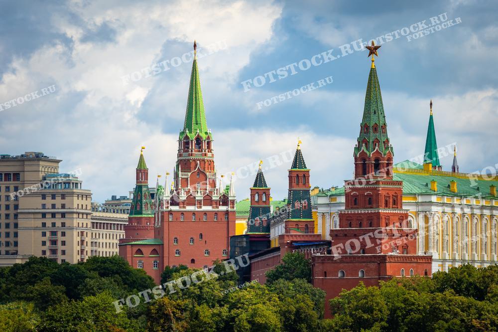 На фотографии изображены несколько башен Московского Кремля с ярко-зелеными шпилями и золотыми звездами. На переднем плане — Никольская и Сенатская башни, символы мощи и истории России. За ними виднеются стены Кремля и купола Большого Кремлевского дворца. Здания окружают густые деревья, что подчеркивает гармонию природы и архитектуры в центре Москвы.