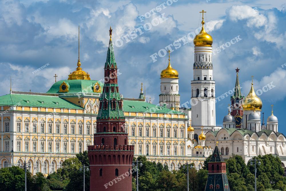 На фотографии изображён Московский Кремль с его величественными зданиями и золотыми куполами. В центре композиции виден Большой Кремлёвский дворец с зелёной крышей и золотым шпилем. Справа расположены купола Успенского собора и колокольня Ивана Великого, которые являются выдающимися памятниками русской архитектуры. На переднем плане выделяется Кремлёвская башня с красным кирпичным фасадом. Голубое небо и облака дополняют красочную композицию.