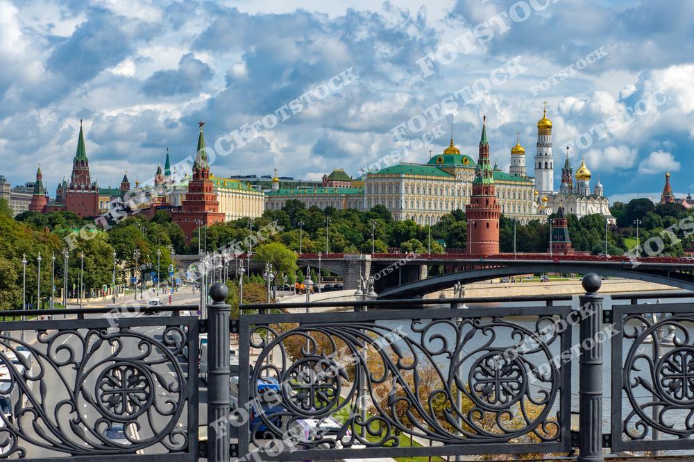 На фотографии изображён Кремль — сердце Москвы и один из главных символов России. В центре видны Кремлёвская стена, башни, а также Большой Кремлёвский дворец с его золотым куполом. Справа возвышаются купола Успенского собора и колокольня Ивана Великого. На переднем плане виден мост через Москва-реку, украшенный изысканными коваными перилами. Летний день с облаками добавляет фотографии насыщенности и подчёркивает красоту этого исторического места.