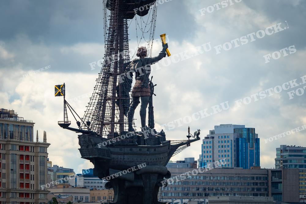 На фотографии изображен памятник Петру I, созданный скульптором Зурабом Церетели, расположенный в Москве. Монумент представляет собой фигуру Петра Великого, стоящего на корабле с поднятыми парусами, что символизирует его реформы и модернизацию Российского флота. Фон изображения показывает современные здания Москвы, подчеркивая контраст между историческим значением памятника и современной архитектурой города. Фотография может быть использована в культурных и исторических публикациях, образовательных материалах и туристических брошюрах.