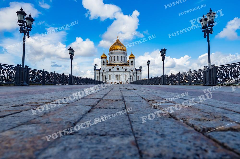 На фотографии изображёнвид на Храм Христа Спасителя в Москве, снятый с уровня мостовой. Золотые купола храма величественно возвышаются над элегантной чугунной оградой моста, украшенной фонарями.  Это изображение может быть использовано в туристических брошюрах, культурных и исторических публикациях, а также в образовательных материалах для иллюстрации русской православной архитектуры.
