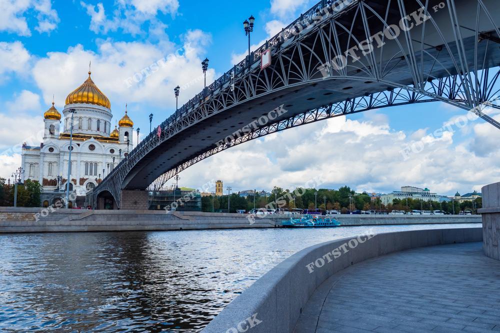 На переднем плане изображён металлический пешеходный мост, соединяющий берега Москва-реки, ведущий к Храму Христа Спасителя. Архитектура моста с изящными перилами гармонирует с величественным видом храма с золотыми куполами. Спокойная поверхность реки отражает голубое небо и лёгкие облака, добавляя снимку яркости и глубины.