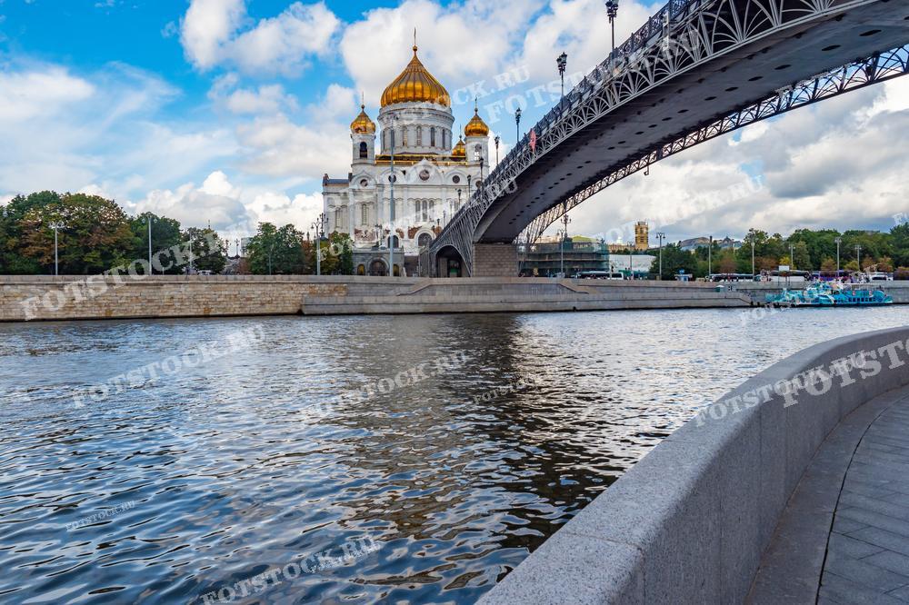 На фотографии изображен величественный Храм Христа Спасителя с его золотыми куполами, возвышающийся над набережной Москвы-реки. Справа виден Патриарший мост, соединяющий берега реки и ведущий к храму. Летний день с голубым небом и облаками придаёт сцене свежесть и яркость. Спокойные воды Москвы-реки отражают мост и храм, добавляя композиции гармонию и спокойствие. Этот вид является одним из самых узнаваемых в Москве.