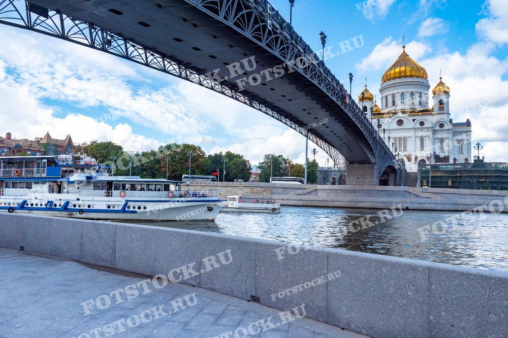 На фотографии изображен вид на Храм Христа Спасителя в Москве, сделанный с набережной реки Москва. Через композицию проходит мост, под которым виден туристический корабль. Здание храма величественно возвышается на фоне городской застройки, подчеркивая его значимость и культурное наследие. Фотография может быть использована в туристических гидах, образовательных материалах, а также в культурных и исторических изданиях для иллюстрации важности этого религиозного сооружения в истории России.