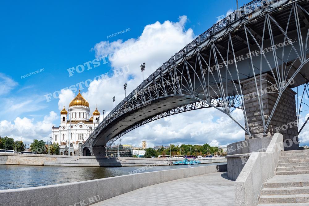 На фотографии изображен Храм Христа Спасителя и металлический мост через Москву-реку. Золотые купола храма сверкают на фоне яркого голубого неба. Летний день в Москве создает атмосферу спокойствия и величия.
