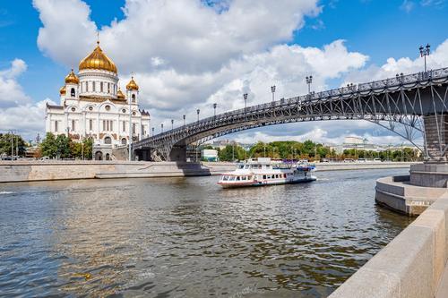 На фотографии изображен живописный вид Москвы в теплый летний день: Храм Христа Спасителя, корабль, плывущий по реке Москве, и Патриарший мост, ведущий к храму. Сцена транслирует спокойствие и красоту русской столицы. Фотография идеально подходит для иллюстрации туристических материалов о Москве, промо-материалов по речным прогулкам и религиозным местам.