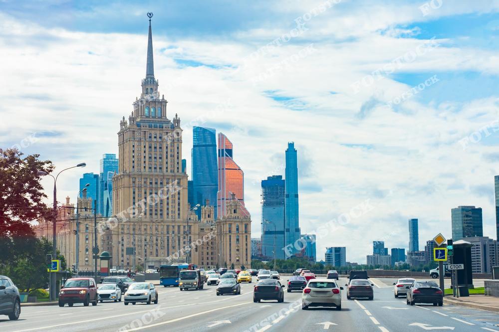 На переднем плане изображена оживлённая магистраль Москвы, ведущая к одной из знаменитых сталинских высоток. Здание, выполненное в стиле сталинского ампира, подчёркивает историческое величие города. На заднем плане возвышаются современные небоскрёбы делового центра Москва-Сити, создавая контраст между архитектурными эпохами. Светлый летний день с ярким небом делает снимок насыщенным, а движение автомобилей придаёт динамику композиции. Это изображение символизирует сочетание истории и современности в столице.