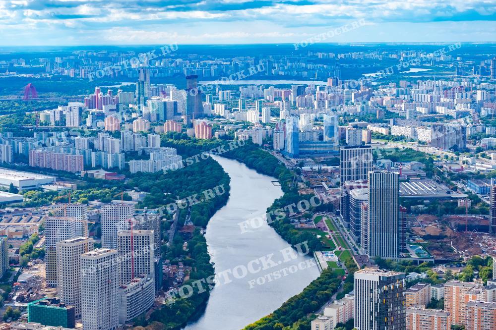 На фотографии представлен панорамный вид на Москву с высоты. В центре снимка протекает Москва-река, огибающая жилые районы с многоэтажными домами и современными зданиями. Видна яркая и разнообразная застройка, окружающая реку, а также зелёные парковые зоны, придающие свежесть урбанистическому пейзажу.