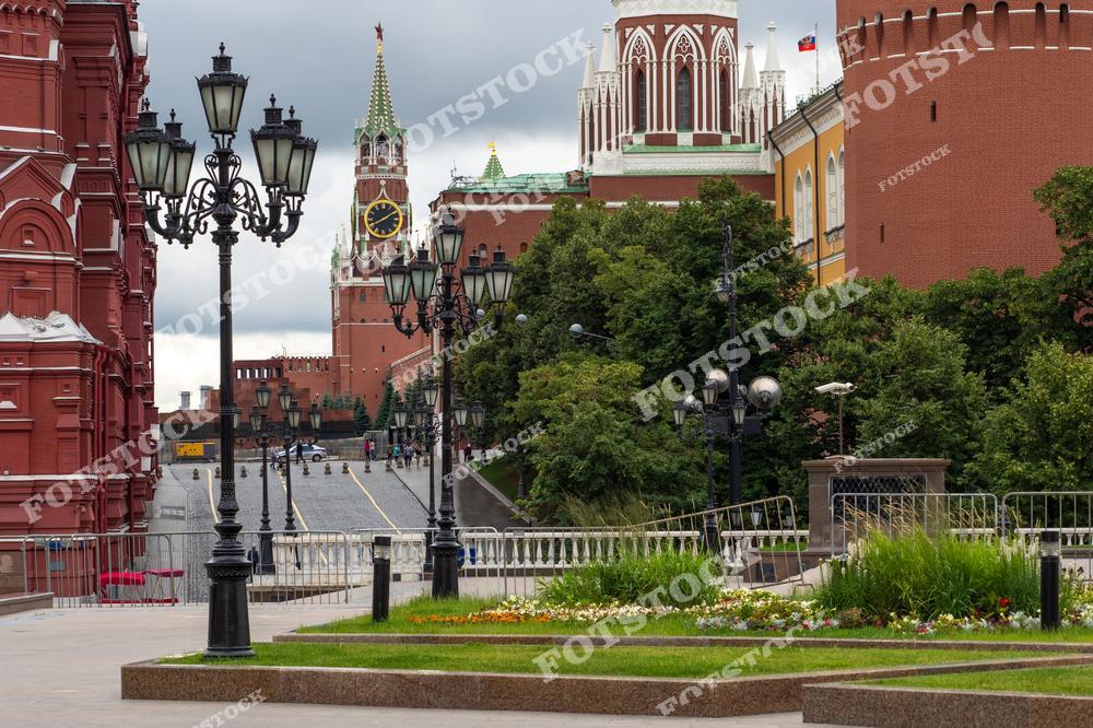 На фотографии запечатлён один из центральных видов Москвы — Спасская башня Кремля с курантами. Слева видны красные стены Исторического музея, справа — части кремлёвской стены и башни. На переднем плане располагается ухоженная зелёная клумба с цветами и уличными фонарями. Вид открывается со стороны Манежной площади, ведущей к Красной. Облачное небо и отсутствие толп придают снимку атмосферу спокойствия. Летняя Москва показана в своём архитектурном величии и историческом духе.