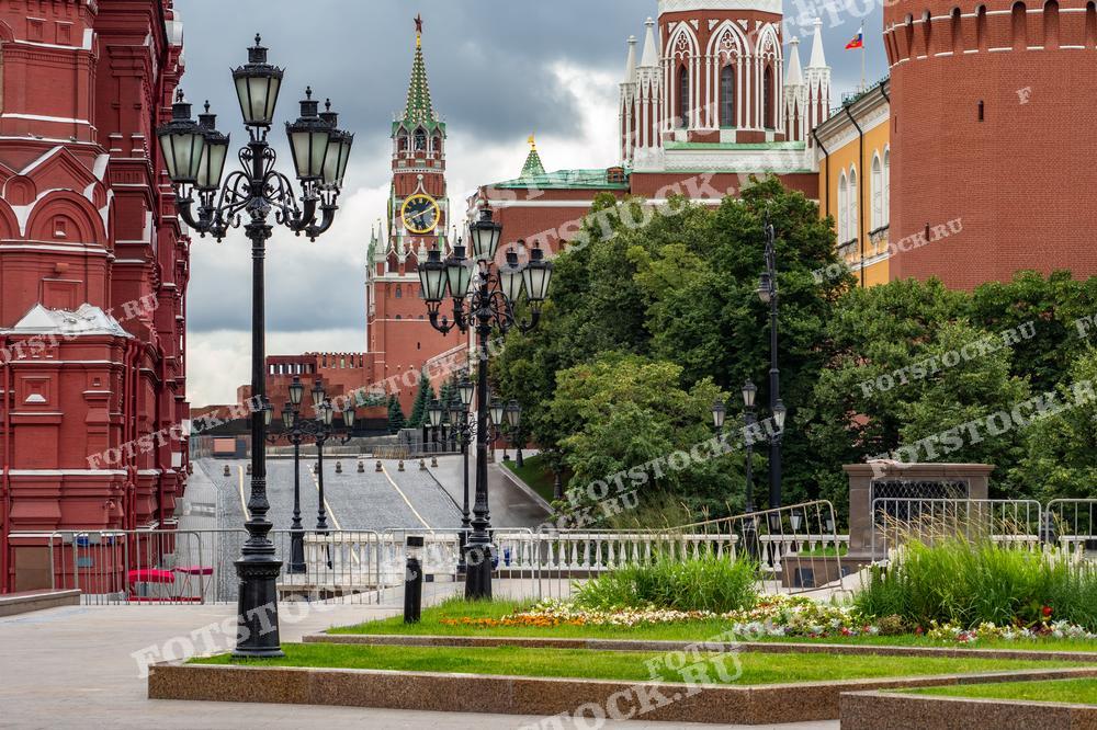 На фотографии изображена часть Красной площади с видом на Спасскую башню Московского Кремля. Ярко-красные стены башни контрастируют с зелеными деревьями и цветочными клумбами на переднем плане. Спасская башня, украшенная знаменитыми курантами, возвышается над площадью.