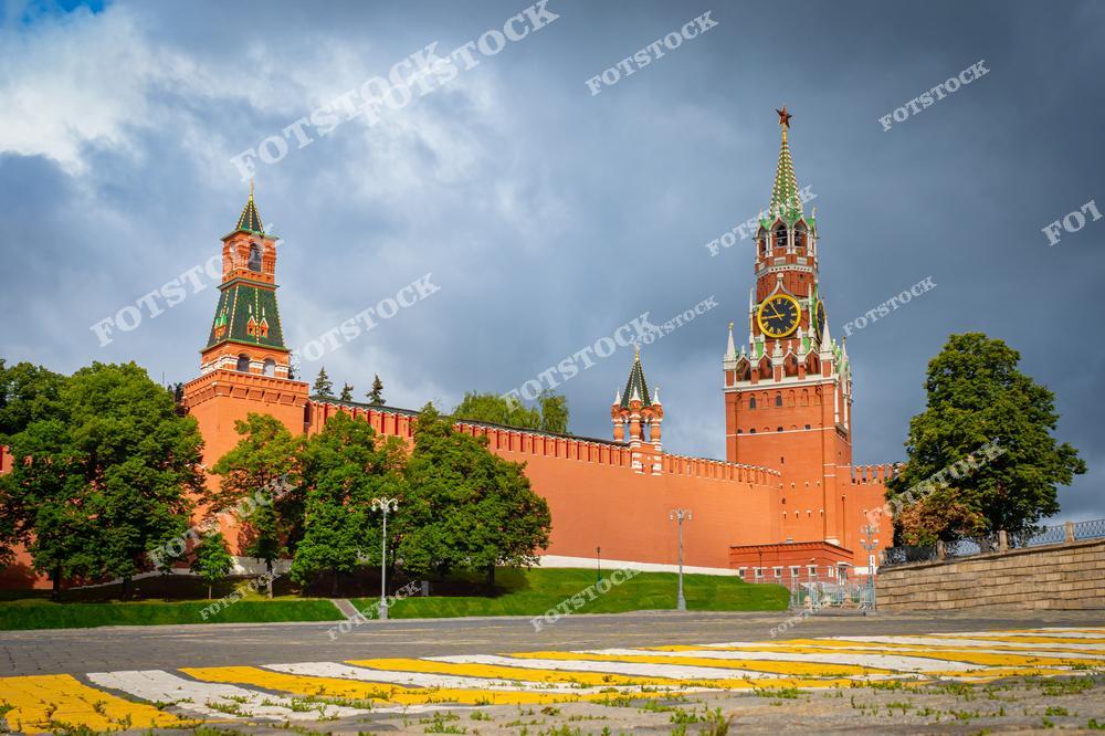 На фотографии изображена часть Кремлёвской стены с башнями — Спасской и Набатной. Спасская башня с курантами, являющаяся символом Москвы, выделяется своим величием и точностью архитектурных форм. Яркое солнечное освещение подчёркивает красный цвет кирпичных стен и зелёные кроны деревьев, окружающих Кремль. На переднем плане видны элементы городской инфраструктуры, включая переход с жёлто-белой разметкой. Темнеющее облачное небо усиливает драматический контраст между светом и тенью, делая композицию более выразительной.