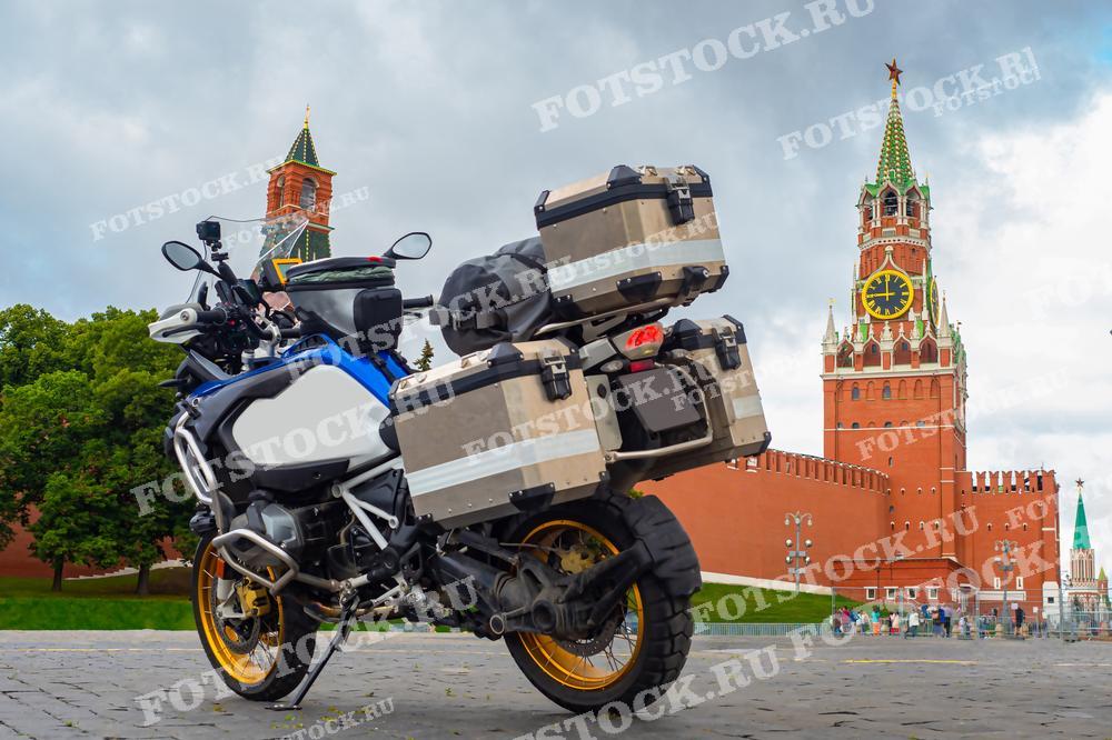 На фотографии изображён туристический мотоцикл с багажными кейсами на фоне Красной площади и Спасской башни Кремля в Москве. Мотоцикл стоит на площади, что символизирует путешествия и приключения в исторических местах города. Спасская башня с часами на фоне ярко выражает историческую значимость и архитектурную красоту места. Это изображение может привлекать внимание любителей мотоциклов и путешественников, а также может быть использовано в туристических и путеводительных материалах.