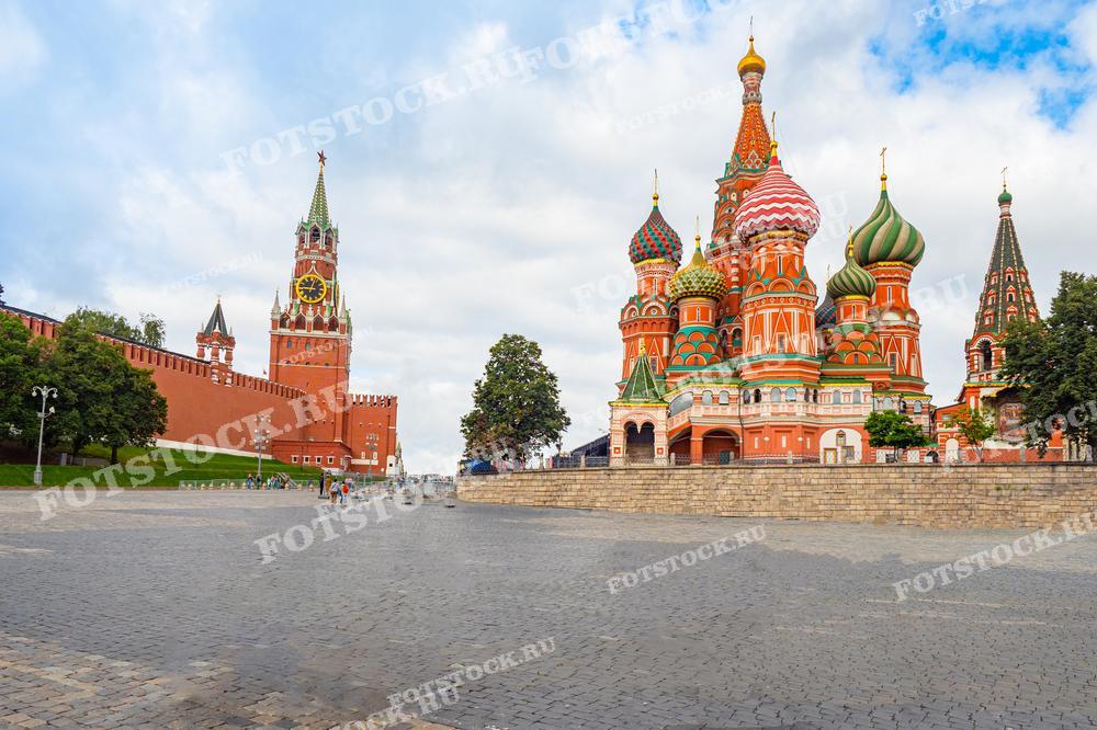 На фотографии изображена Красная площадь в Москве с видом на Спасскую башню Кремля и собор Василия Блаженного. На переднем плане вымощенная площадь, на заднем плане башня с часами и красочная архитектура собора. Летний день с голубым небом и облаками создает яркую и праздничную атмосферу.