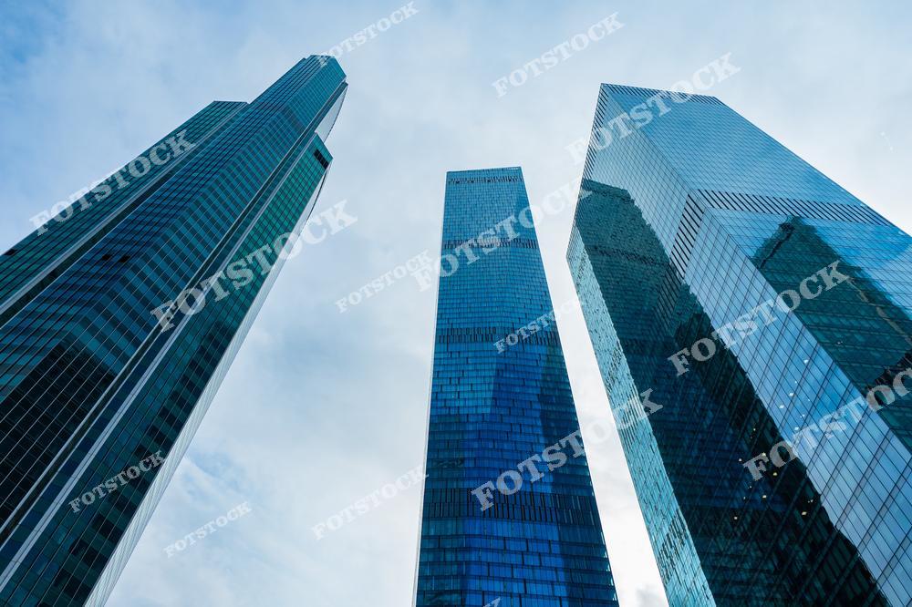 На фотографии изображены три высотных здания комплекса Москва-Сити, снятые с нижнего ракурса. Их стеклянные фасады отражают свет неба, создавая впечатление прозрачности и лёгкости. Архитектурный стиль небоскрёбов подчёркивает современные технологии и инновации. Контраст между масштабностью зданий и мягкостью облаков создаёт гармоничную композицию. Москва-Сити символизирует прогресс и экономическую мощь российской столицы.