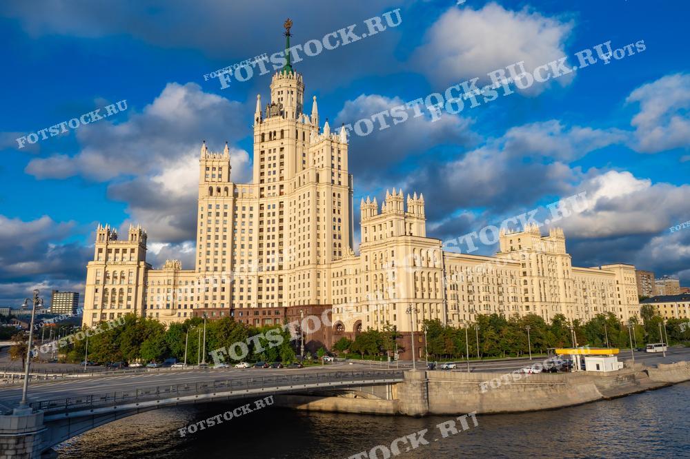 На фотографии изображена знаменитая сталинская высотка на Котельнической набережной — одно из самых знаковых зданий Москвы. Это монументальное строение, построенное в стиле сталинского ампира, является символом советской архитектуры. Его высокие башни и шпиль венчают композицию, возвышаясь над Москвой-рекой. Внизу виден мост, ведущий через реку, и зелёные деревья вдоль набережной. Яркий солнечный свет и облачное небо подчеркивают величие этого архитектурного шедевра.