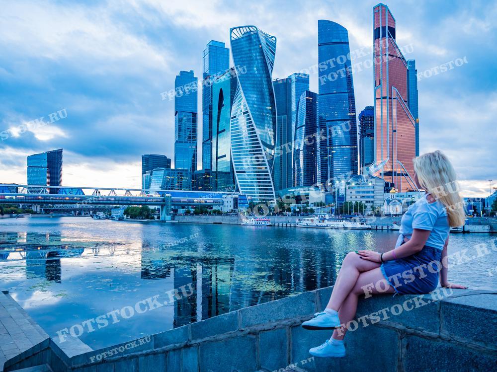 На фотографии изображена молодая женщина, сидящая на набережной и смотрящая на панораму Москва-Сити. За спиной у женщины видны знаковые здания делового района, отражающиеся в водах реки. Это изображение может использоваться в материалах, посвященных городскому пейзажу или современной жизни в Москве.