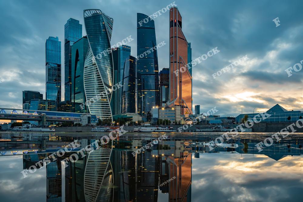 На снимке изображен деловой район Москва-Сити, известный своими современными небоскребами. Башни комплекса отражаются в спокойной поверхности реки Москвы, создавая эффект зеркальности.