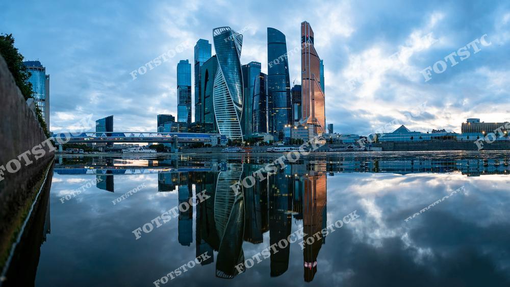 На фотографии запечатлён деловой центр Москвы — Москва-Сити, отражающийся в гладкой поверхности Москва-реки. Комплекс состоит из современных небоскрёбов, включая башни «Эволюция», «Федерация» и другие знаковые здания. Время съёмки — раннее утро, когда город только начинает оживать, а небо затянуто лёгкими облаками. Арочная пешеходная галерея на заднем плане добавляет футуристичности сцене. Москва-Сити является символом современной архитектуры и финансового центра столицы. Общая атмосфера кадра — спокойствие и архитектурное величие.