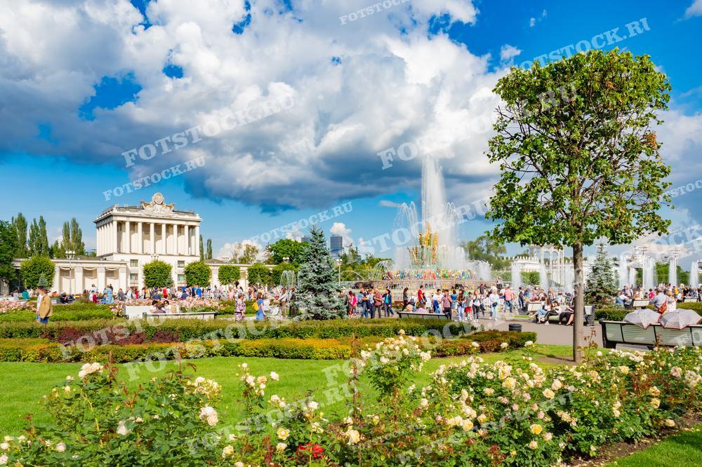 На фотографии изображен фонтан "Каменный цветок" на ВДНХ в Москве в летний день. Вокруг фонтана прогуливаются люди, на переднем плане видны цветущие клумбы. Фото можно использовать для иллюстраций, рекламы и туристических материалов.
