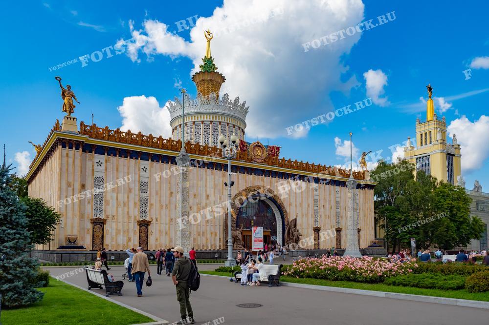 На фотографии изображен Центр славянской письменности "Слово" (бывший павильон "Украина") на ВДНХ в Москве в солнечный летний день. Люди прогуливаются и отдыхают возле павильона, на переднем плане видны цветущие клумбы. Фото можно использовать для иллюстраций, рекламы и туристических материалов.