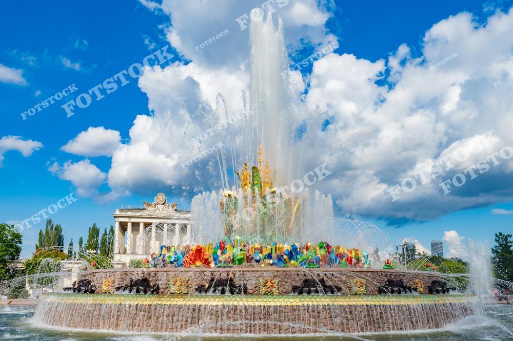 На фотографии изображен фонтан "Каменный цветок", расположенный на ВДНХ в Москве. Этот уникальный архитектурный объект создан в стиле сталинского ампира и украшен яркой мозаикой, символизирующей богатства народов СССР. В центре композиции видны струи воды, которые поднимаются ввысь, создавая ощущение свежести и динамики. На заднем плане заметна часть павильонов выставочного центра, гармонично дополняющих общий пейзаж. Летнее голубое небо с облаками подчеркивает яркость и красоту фонтана.