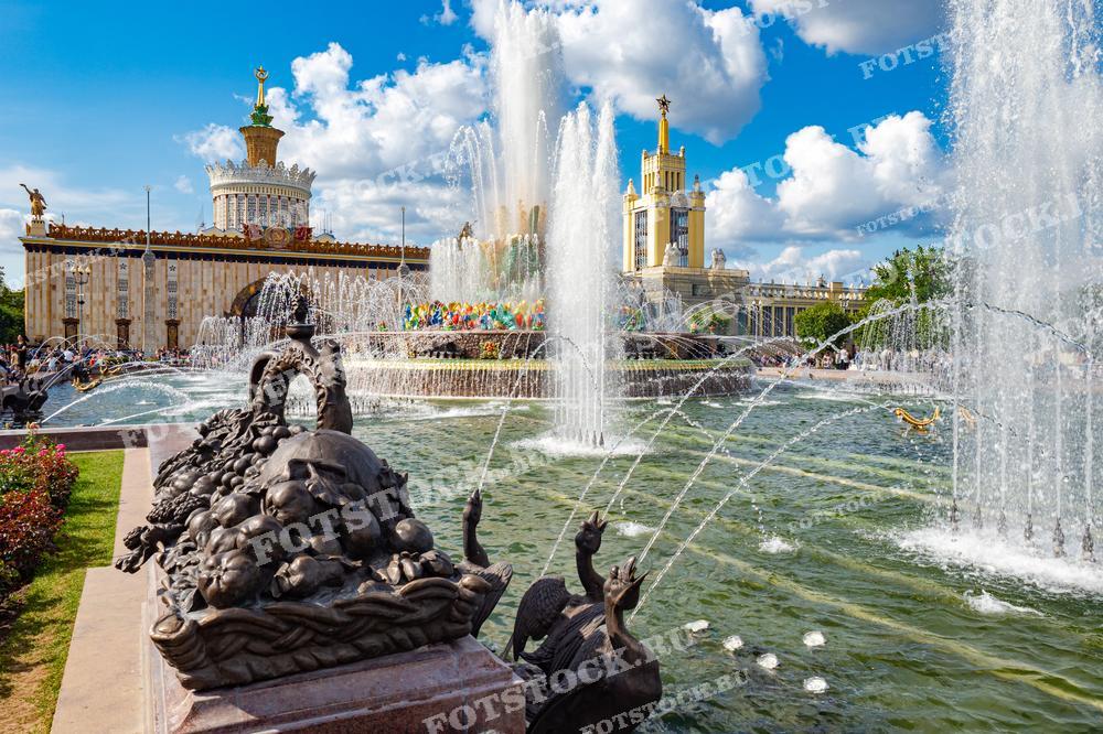 На фотографии изображены фонтаны ВДНХ в Москве, на заднем плане видны павильоны с характерной советской архитектурой. Фонтан "Каменный цветок" и другие элементы украшают территорию выставки в солнечный день.