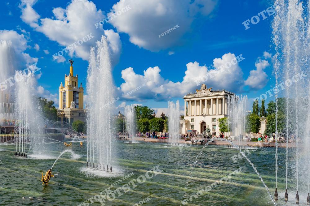 На фотографии изображен знаменитый фонтан "Дружба народов", расположенный на территории ВДНХ в Москве. Вода из многочисленных струй создает эффект торжественности и гармонии. На заднем плане видны архитектурные павильоны выставки, выполненные в стиле сталинского ампира. Золотые детали и летний пейзаж делают композицию особенно яркой и запоминающейся. Посетители, прогуливающиеся вокруг, добавляют жизни и динамики этой сцене.