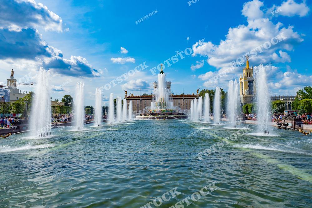 На снимке изображен фонтан "Дружба народов", один из главных символов ВДНХ в Москве. Центральный элемент композиции — золотые скульптуры, символизирующие республики СССР, окружены мощными струями воды. Вокруг фонтана видны павильоны выставочного комплекса. Яркое летнее солнце и голубое небо создают праздничную атмосферу. Гости выставки отдыхают у воды, наслаждаясь живописным ансамблем.