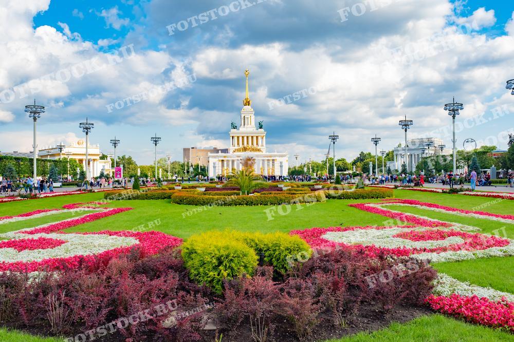 На фотографии изображена центральная аллея ВДНХ в Москве с цветочными клумбами и павильонами на заднем плане в солнечный летний день. Люди прогуливаются и отдыхают на территории выставки. Фото можно использовать для иллюстраций, рекламы и туристических материалов.