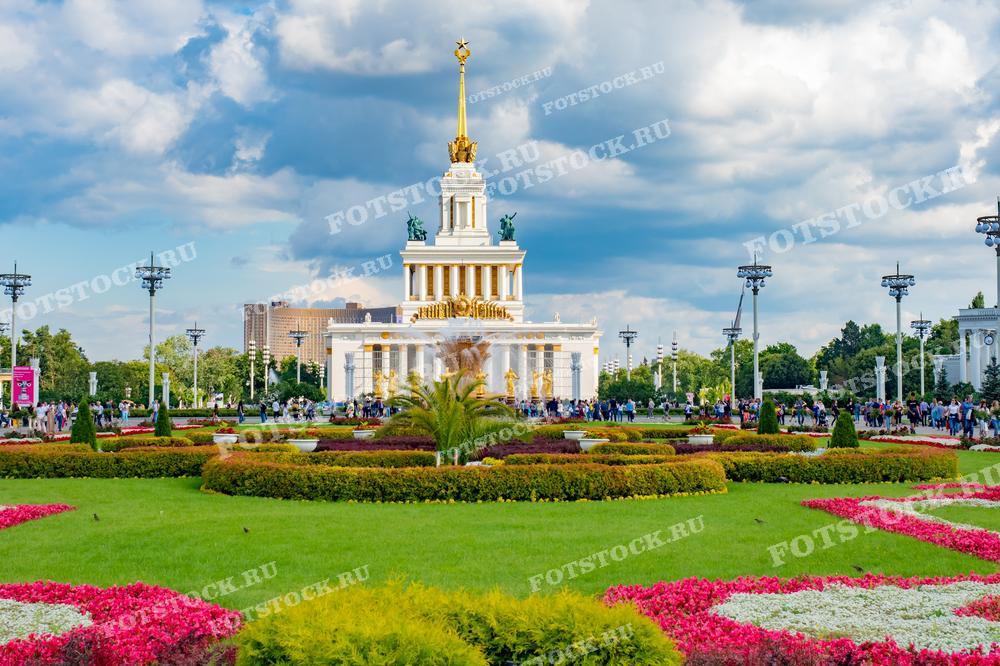 На фотографии изображен главный павильон ВДНХ в Москве с пышными цветниками на переднем плане. Здание украшено золотым статуарным декором, а его архитектура в стиле соцреализма отражает культурное наследие Советского Союза. Этот вид особенно красив благодаря облакам на фоне, которые добавляют драматизма композиции. Идеально подходит для иллюстративного материала о ВДНХ как культурном и историческом объекте в России.